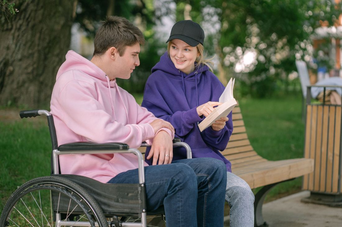 A support worker and a man in a wheelchair reading a book together on a park bench